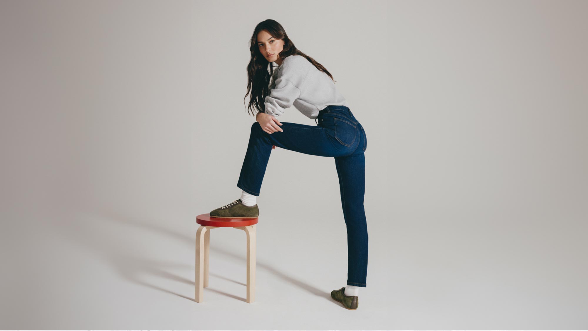 A person in a casual outfit with a gray sweater and jeans poses with a foot on a stool against a plain background.