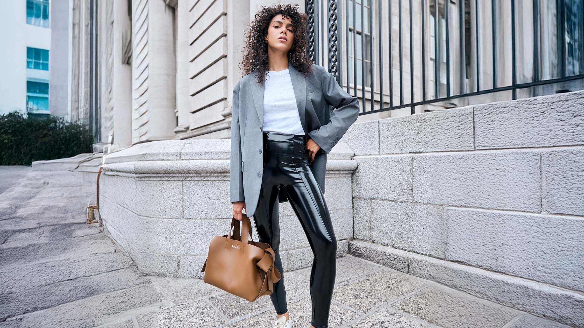 A woman in a blazer, white shirt, and shiny Spanx leggings, holding a brown handbag, poses confidently on a city street.