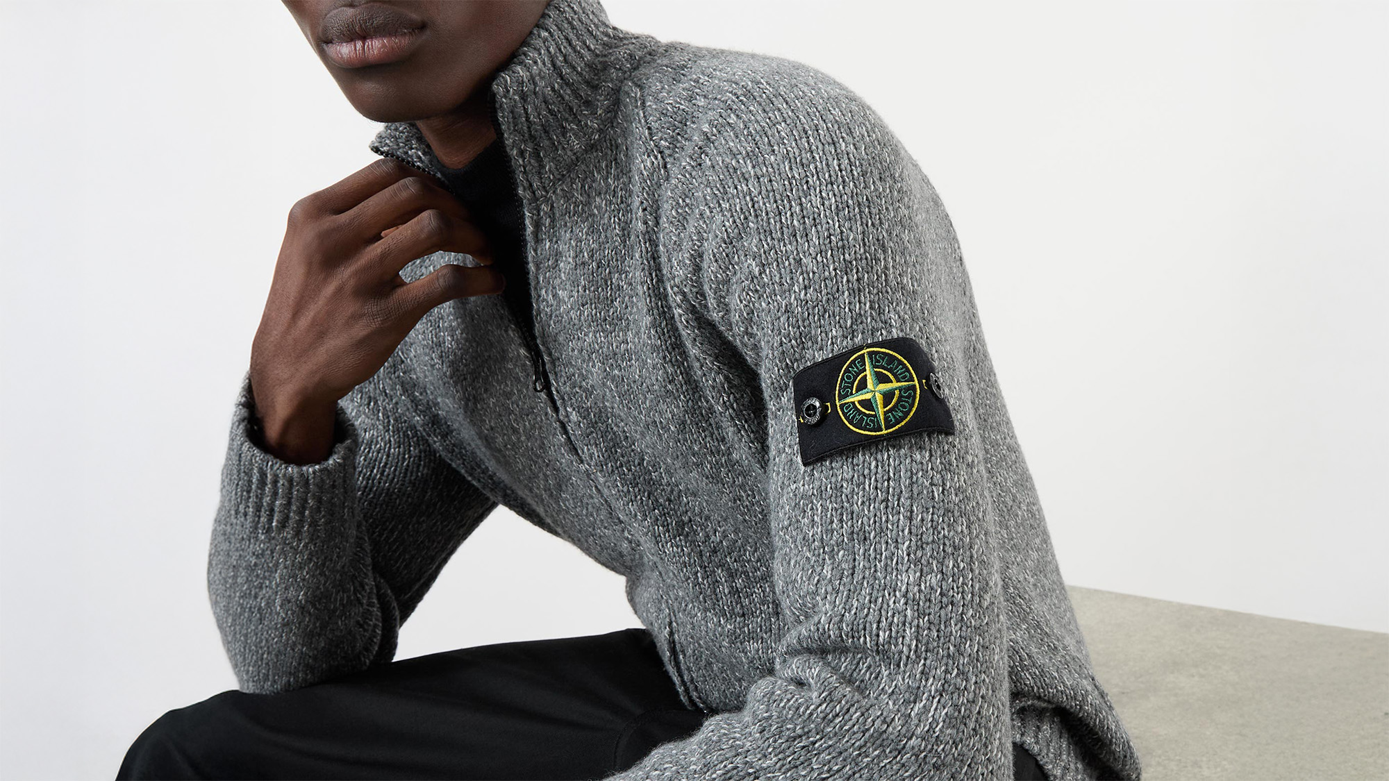 A man wearing a grey knitted Stone Island jumper sits.