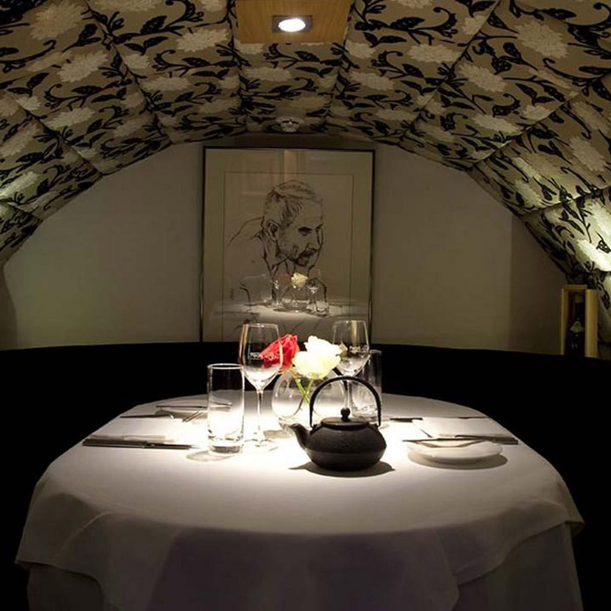 Elegant dining table with white tablecloth, wine glasses, and a floral centerpiece in a dimly lit room with patterned ceiling and artwork.