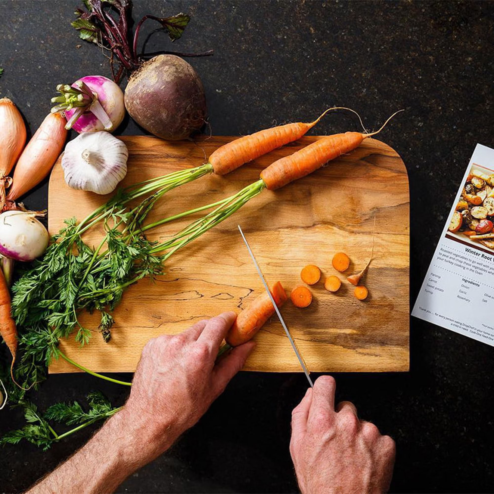 A person slices carrots on a wooden cutting board surrounded by various vegetables, with a recipe card nearby.