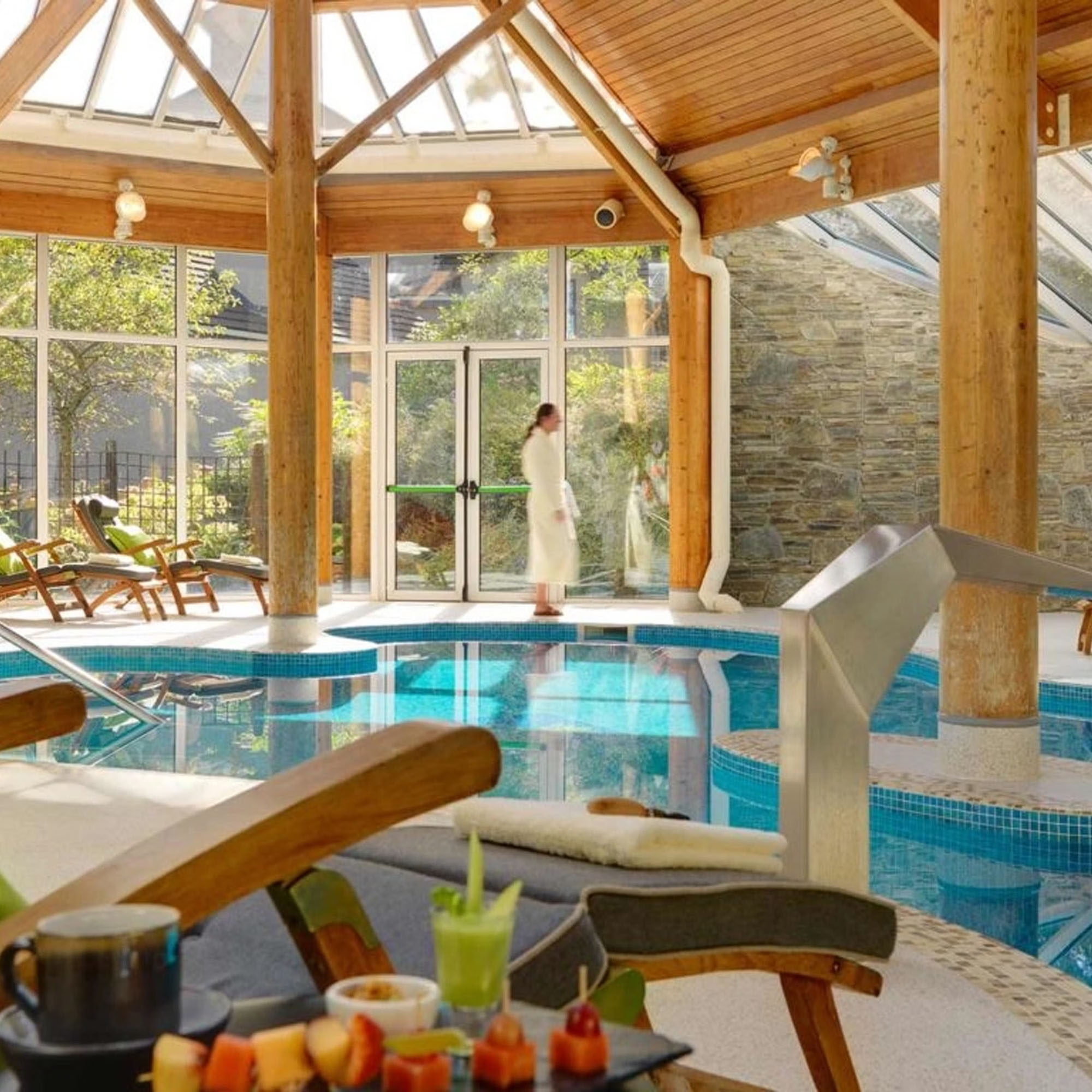 A serene indoor pool area with wooden beams and a glass ceiling. A person in a robe walks by, and there's a table with drinks and snacks.