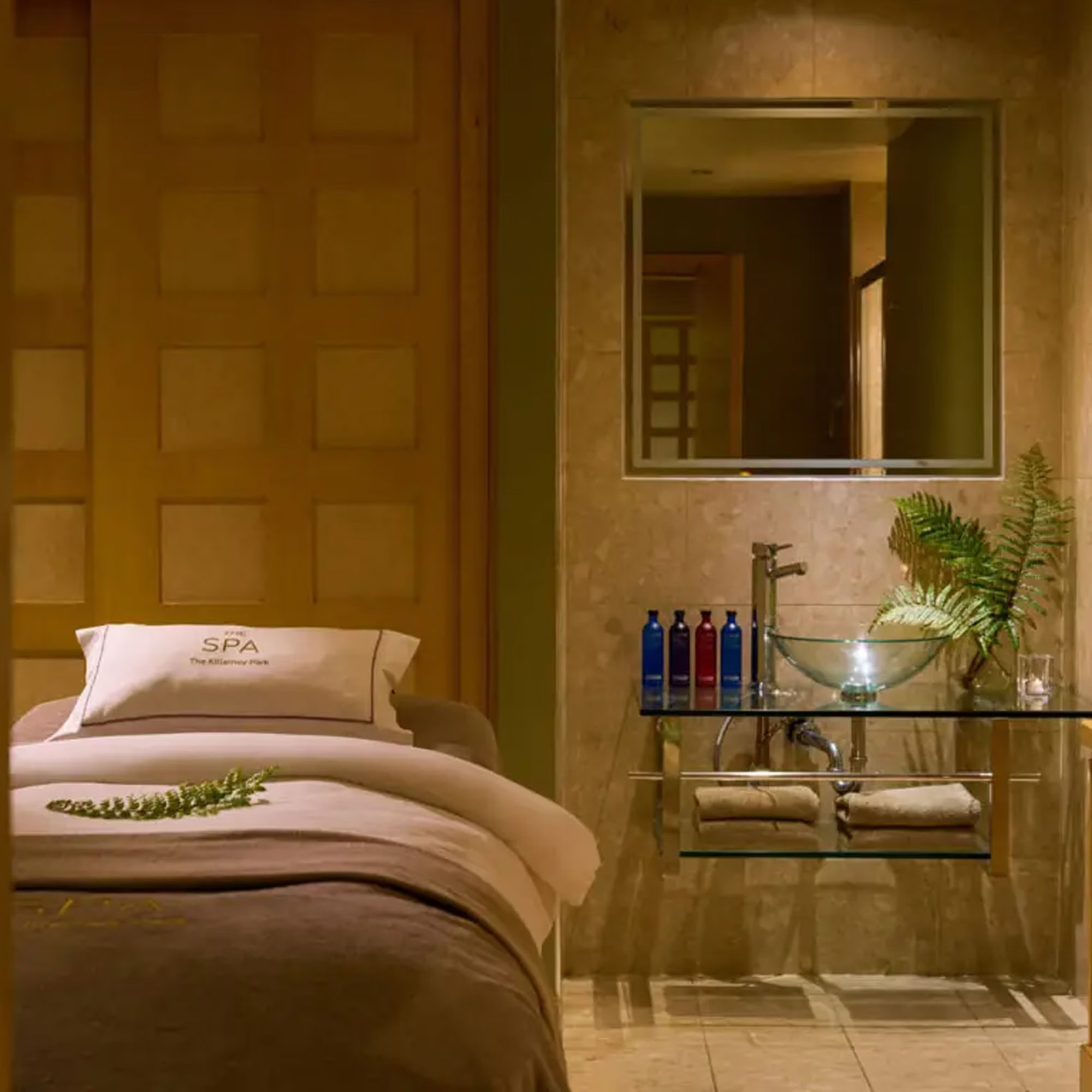 A serene spa room with a massage table, neatly placed towels, various bottles, and decorative plants on a glass shelf.