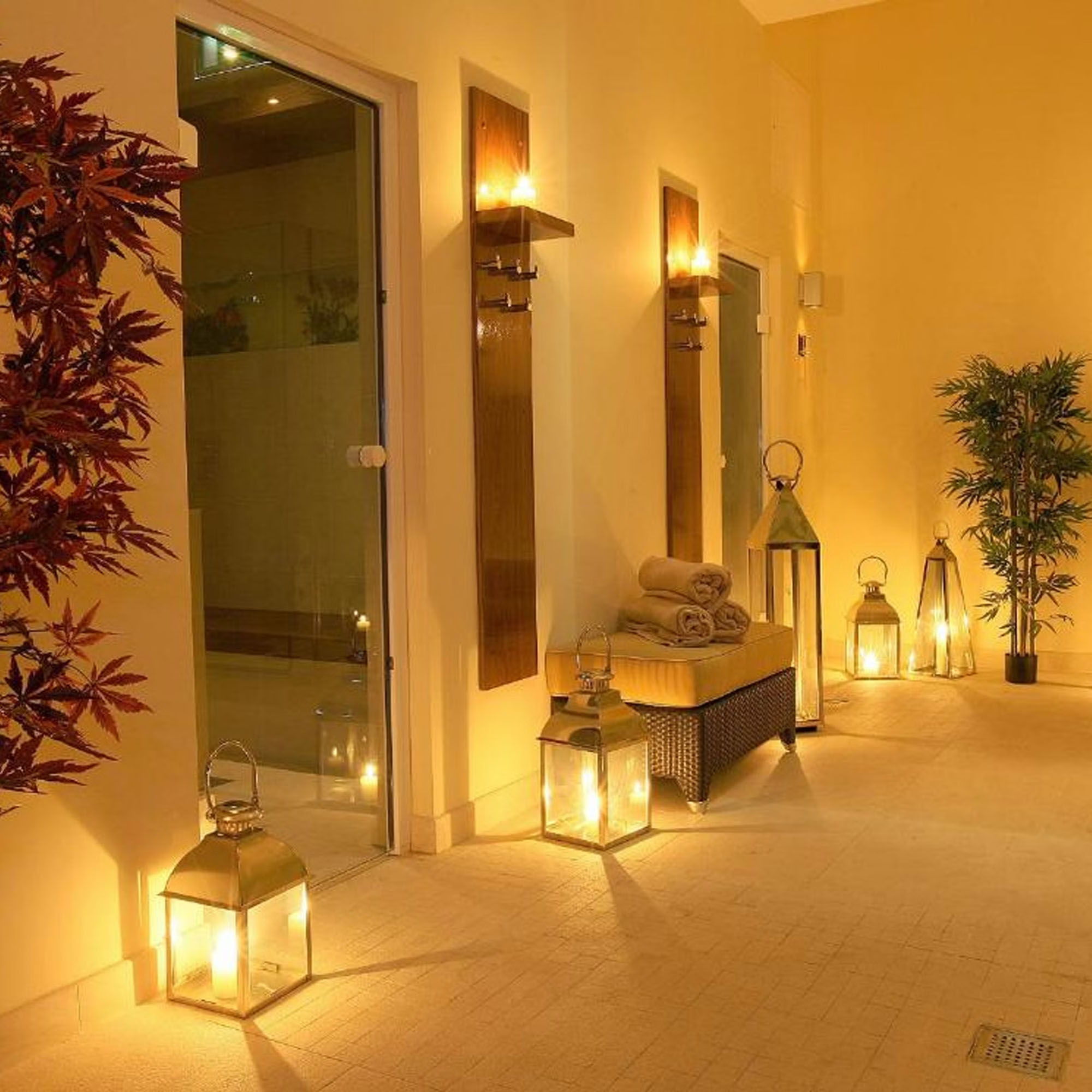 A warmly lit spa hallway with lanterns, a potted plant, rolled towels, and a glass door.