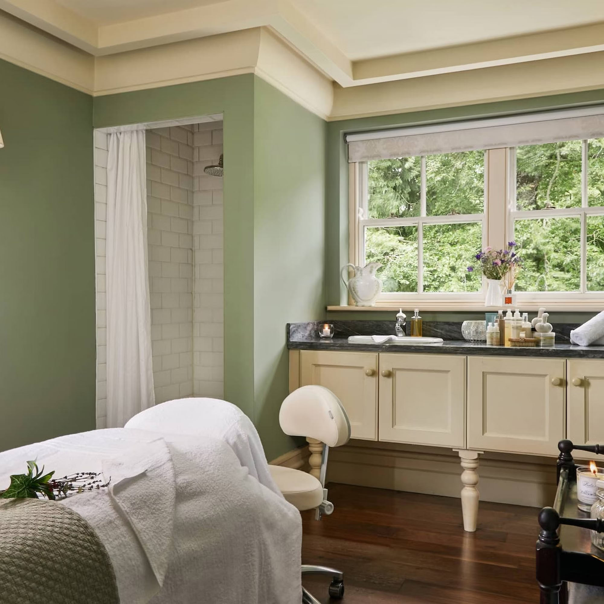 A serene bathroom with green walls, a large window, white cabinets, and a shower with a curtain. There's a treatment table in the foreground.