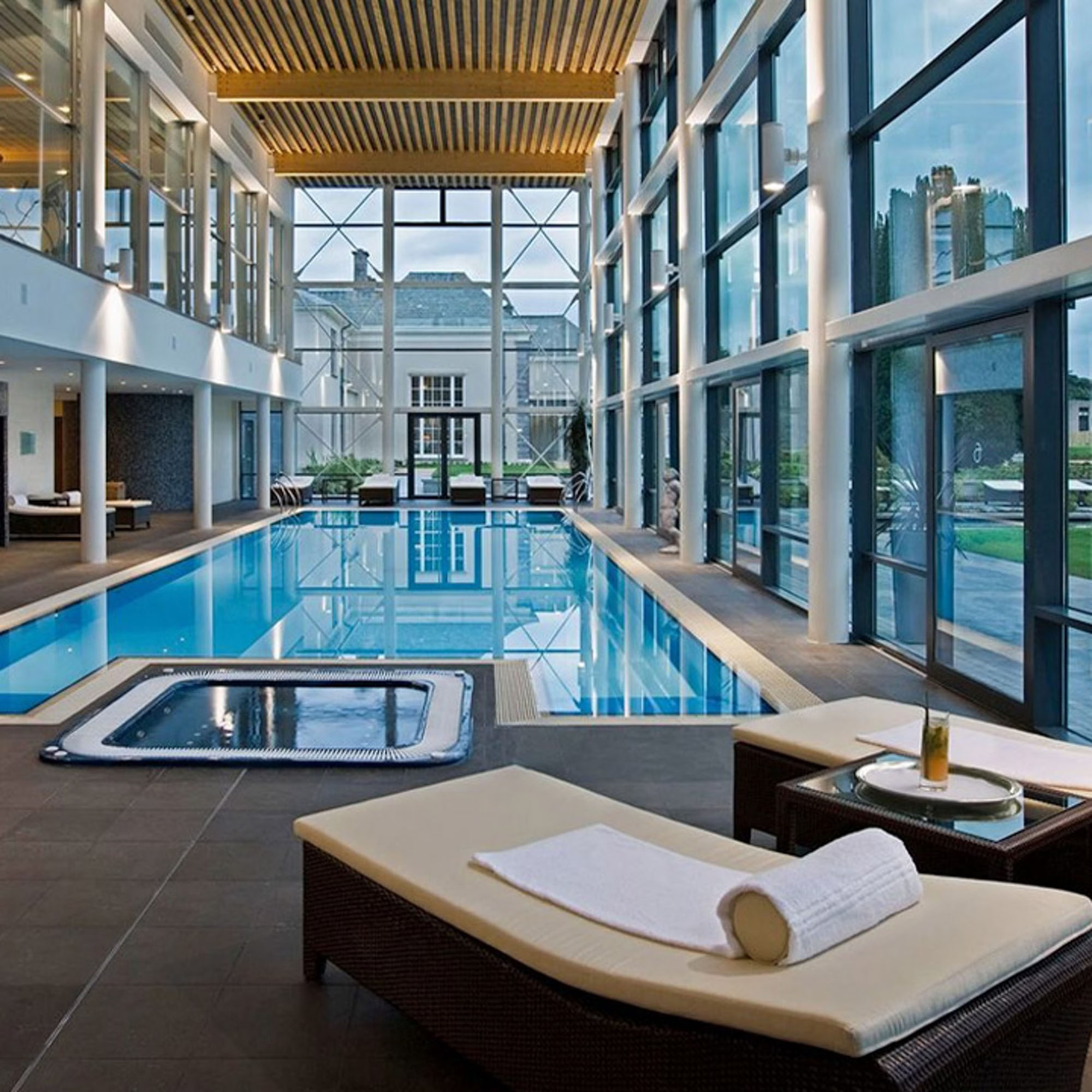 Indoor pool area with large windows, a hot tub, lounge chairs, and a towel. A bottle and glass are on a small table nearby.