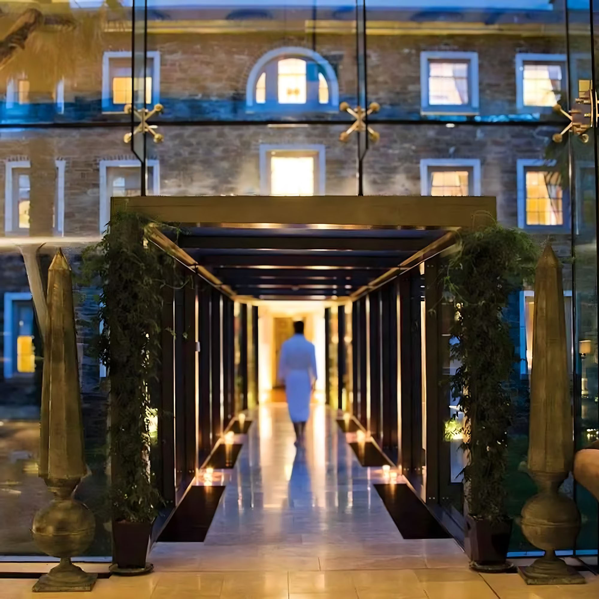 A person in a white robe walks through a glass corridor adorned with plants, facing a lit-up brick building outside.