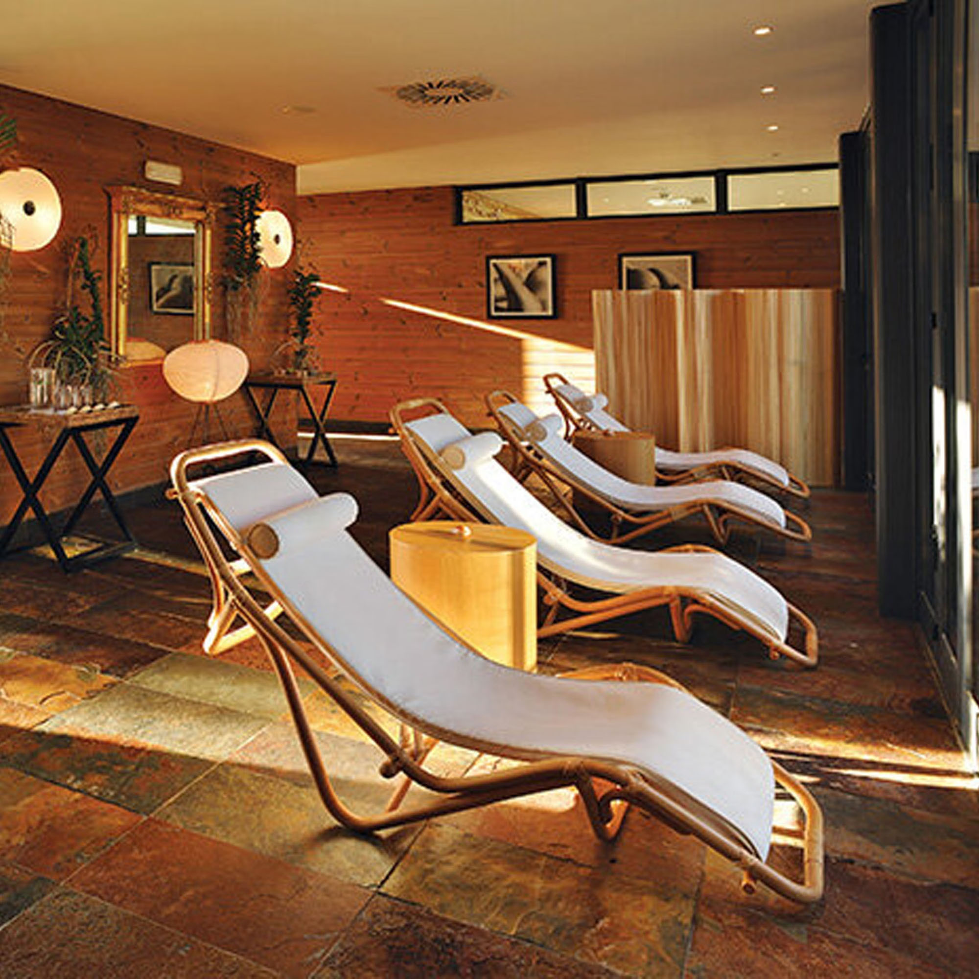 Modern lounge area with wooden deck chairs, soft cushions, warm lighting, and potted plants.