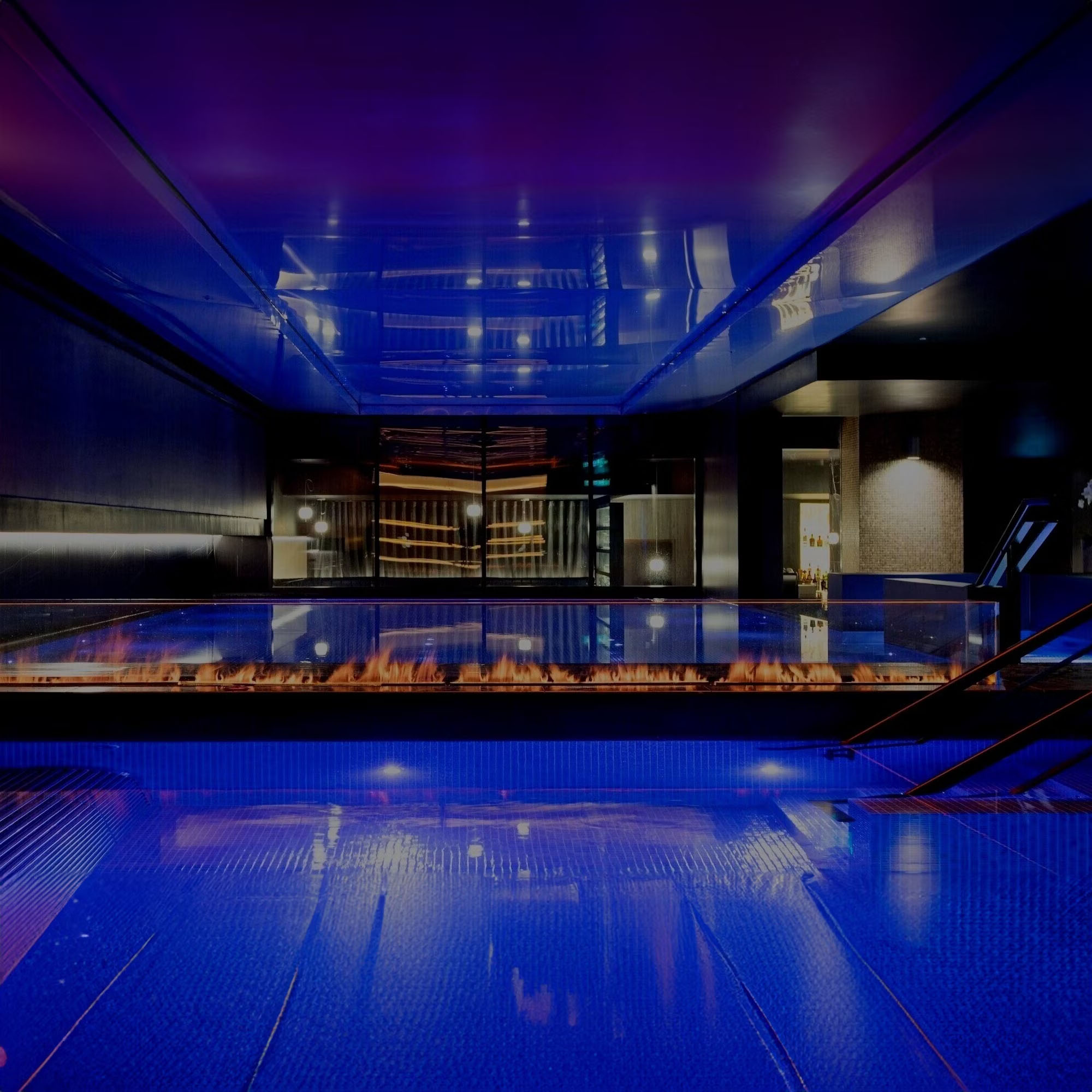 Modern, dimly lit indoor pool with blue and purple lighting, featuring reflective surfaces and sleek design elements.