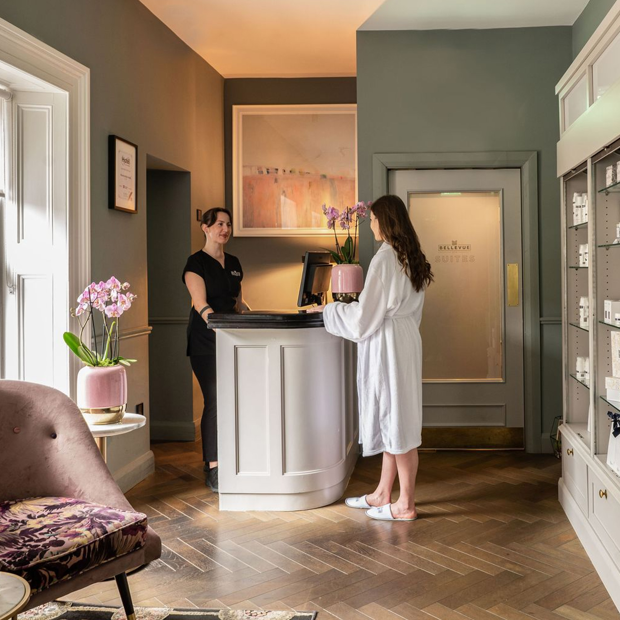 A woman in a robe stands at a reception desk talking to a staff member in a spa-like setting.