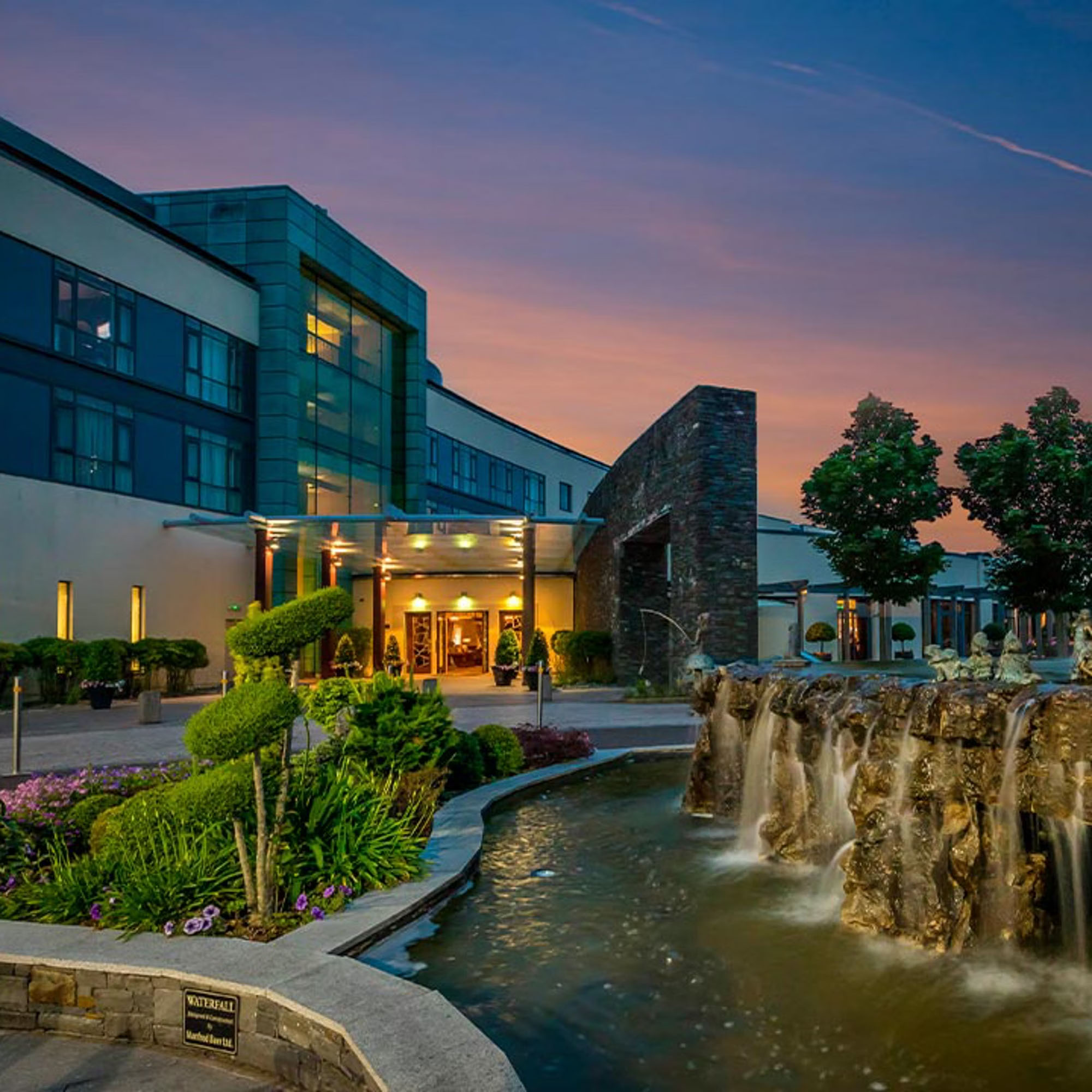 Modern building with lit entrance, surrounded by greenery and a stone waterfall, at sunset.