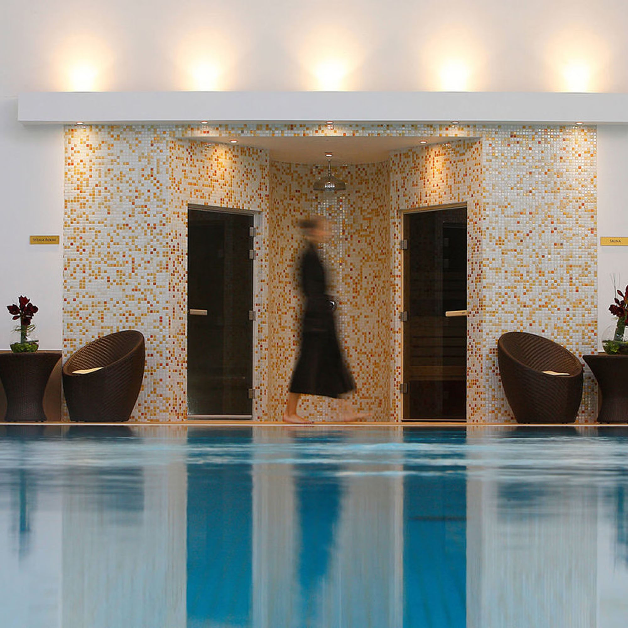 A person in a robe walks past sauna doors near an indoor pool, with warm lighting and decorative chairs.