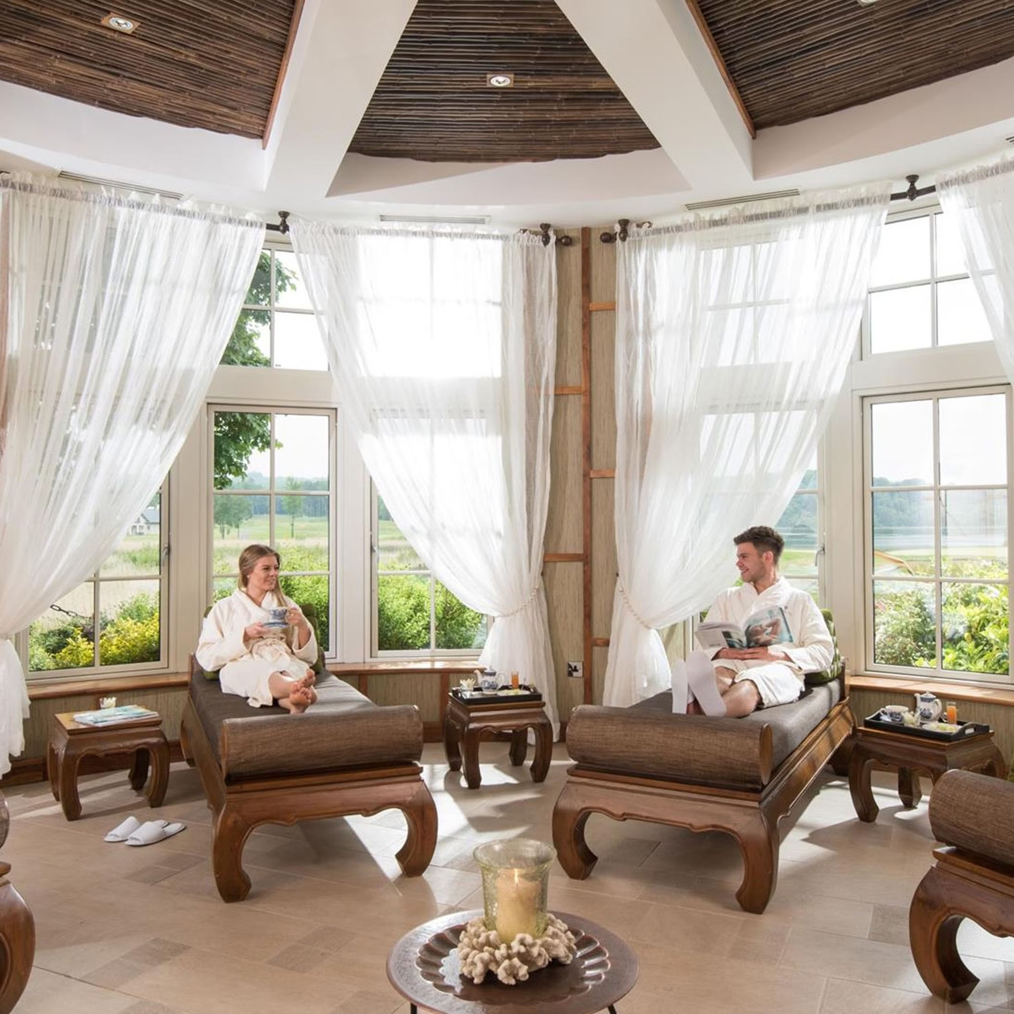 A serene spa room with two people relaxing on lounge chairs, surrounded by sheer drapes and large windows overlooking greenery.