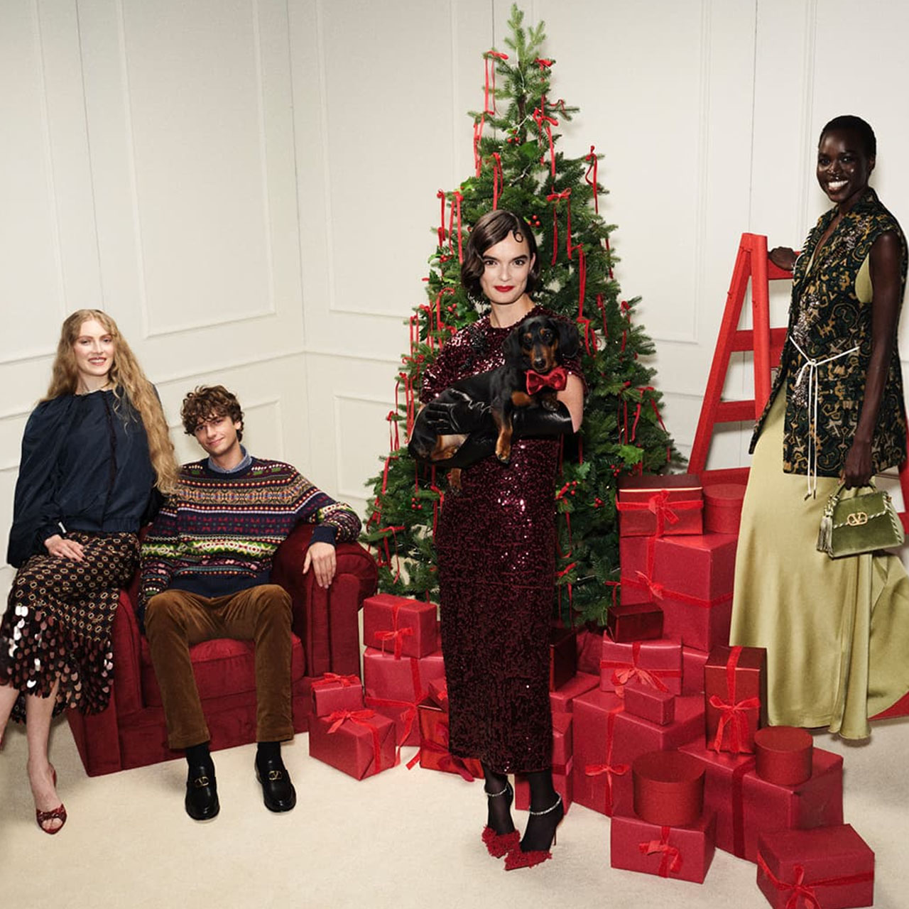 Four people dressed stylishly pose by a decorated Christmas tree with red gift boxes.