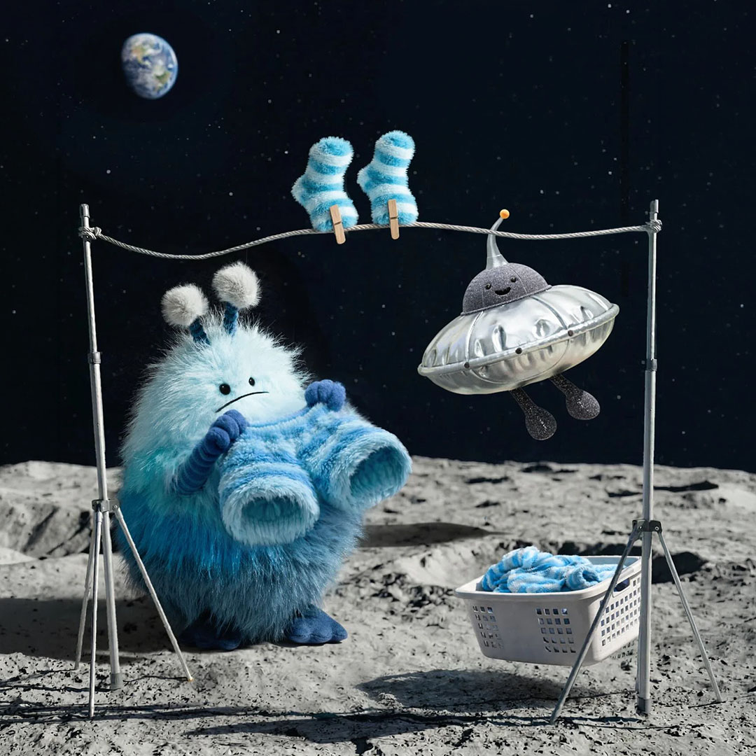 A blue furry creature hangs socks on a line beside a UFO on a lunar surface, with Earth visible in the background.