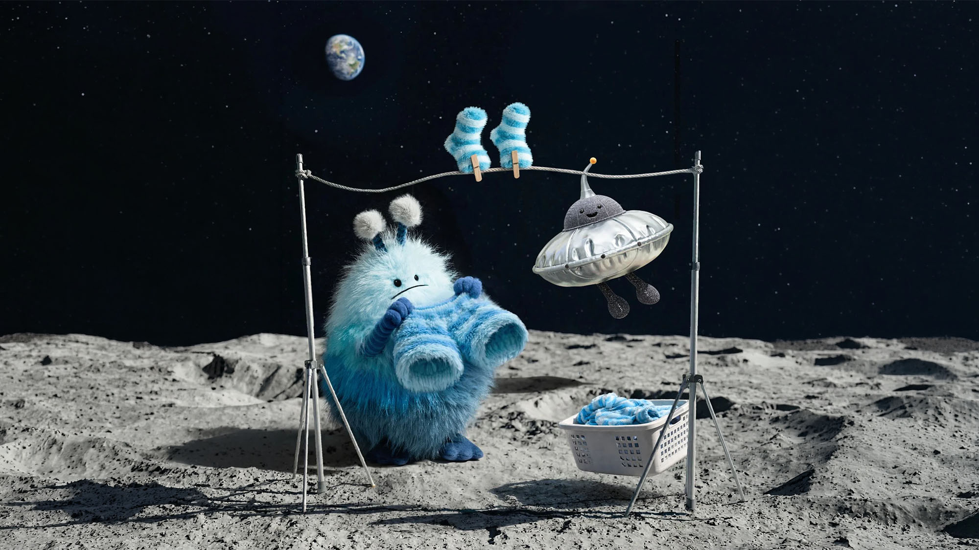 A fluffy blue creature on the moon hangs out laundry alongside a small UFO. Earth and two worm-like beings are in the background.