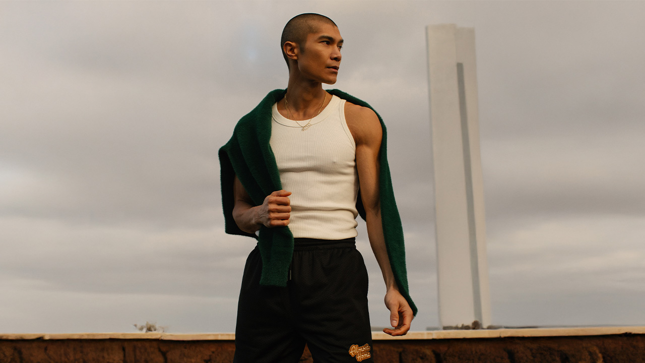 Man in a white tank top and black shorts with a green sweater draped over shoulders, gazing to the side.
