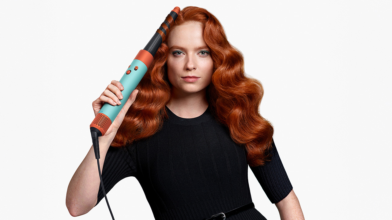 A woman with red hair uses a turquoise curling iron on her curls, wearing a black dress against a plain background.