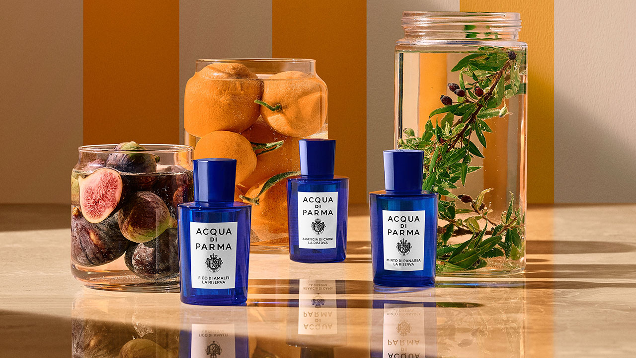 Three Acqua di Parma bottles are displayed with jars of figs, oranges, and herbs, set against an orange and white striped background.