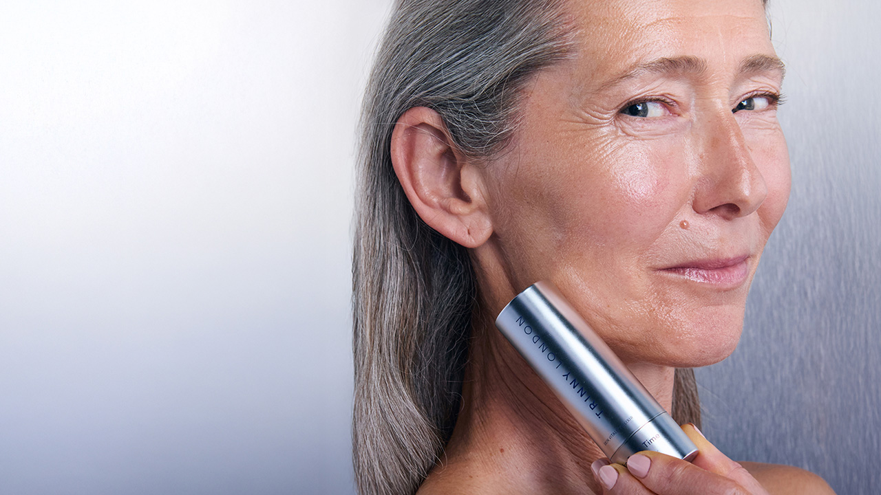 A woman holds a Trinny London product near her face, showcasing smooth skin.