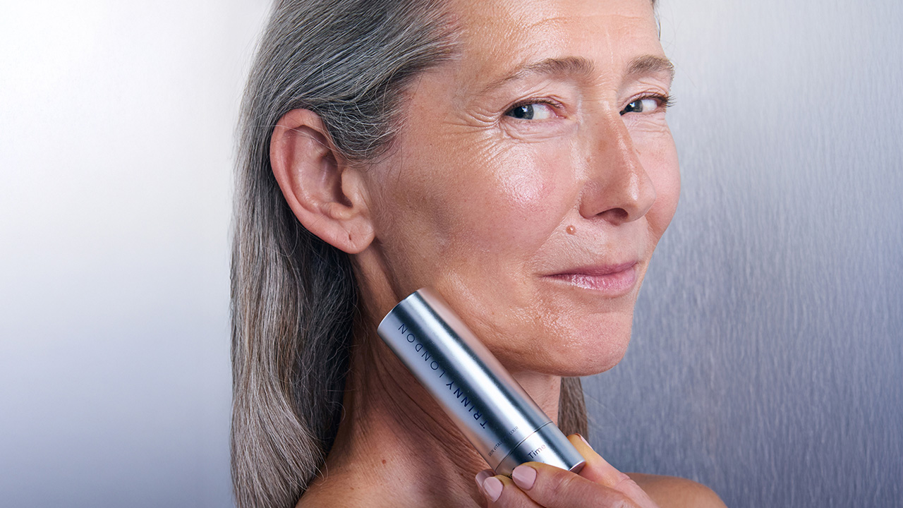 A woman holds a Trinny London product near her face, showcasing smooth skin.