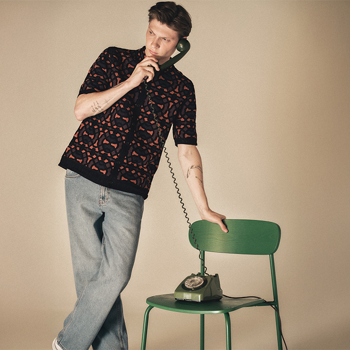 Man in patterned shirt leans on a green chair, talking on a vintage green rotary phone.
