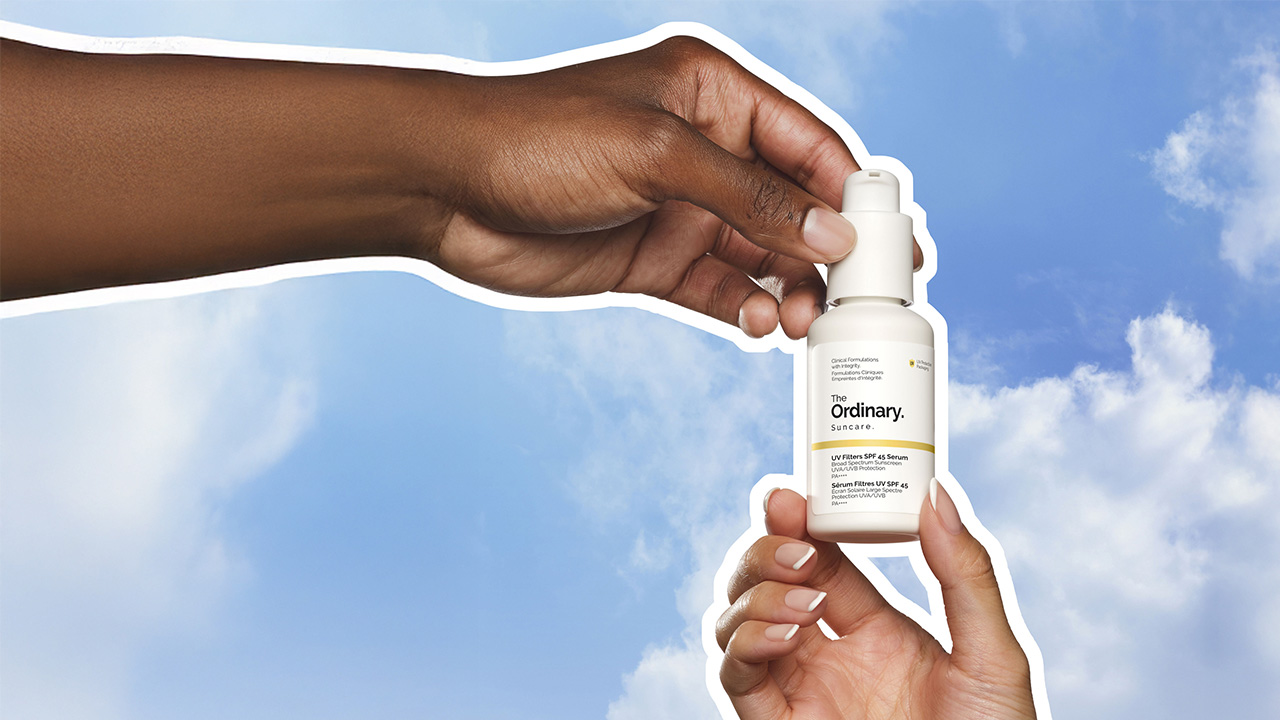Two hands holding 'The Ordinary' sunscreen bottle against a blue sky backdrop.
