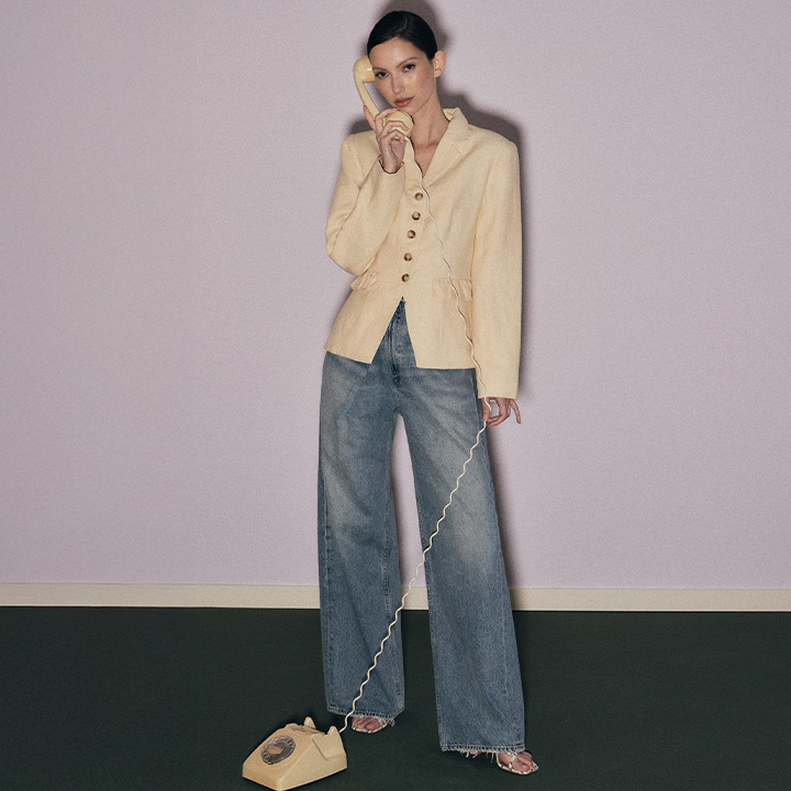 Person in a beige blazer and wide jeans talks on a vintage corded phone in a minimalist room.