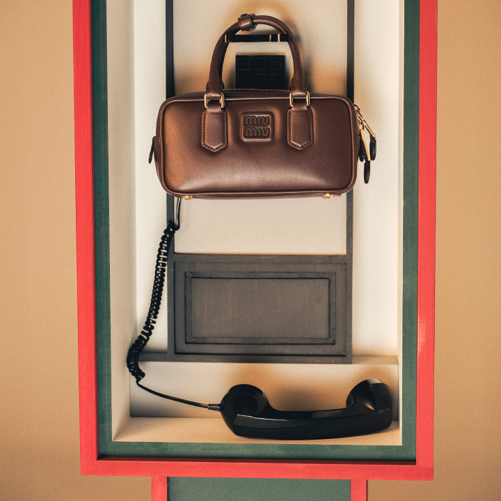 A brown Miu Miu handbag is placed inside an old-fashioned phone booth with a black handset hanging below.