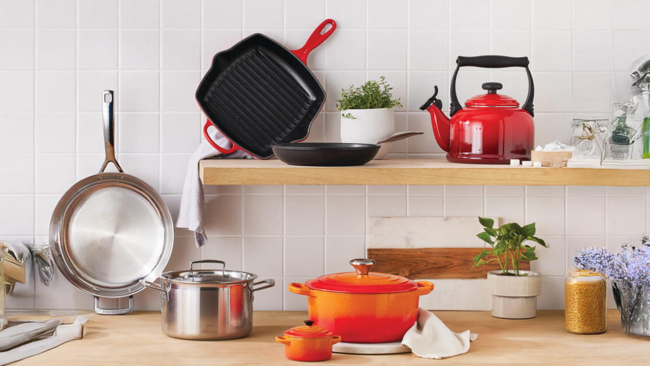 Kitchen scene with Le Creuset cookware: red kettle, grill pan, Dutch oven, stainless steel pot, and small plants on shelves.