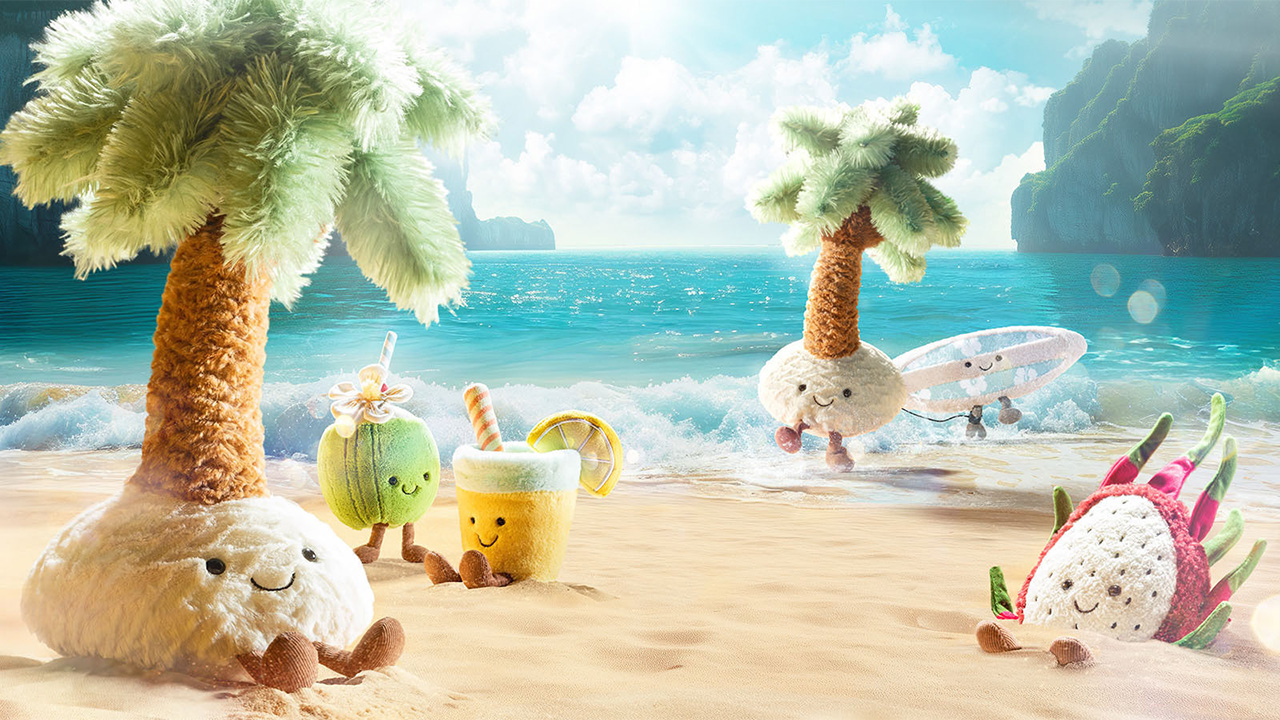 Plush Jellycat toys on a sunny beach, including palm trees and cute fruits, with an ocean backdrop.