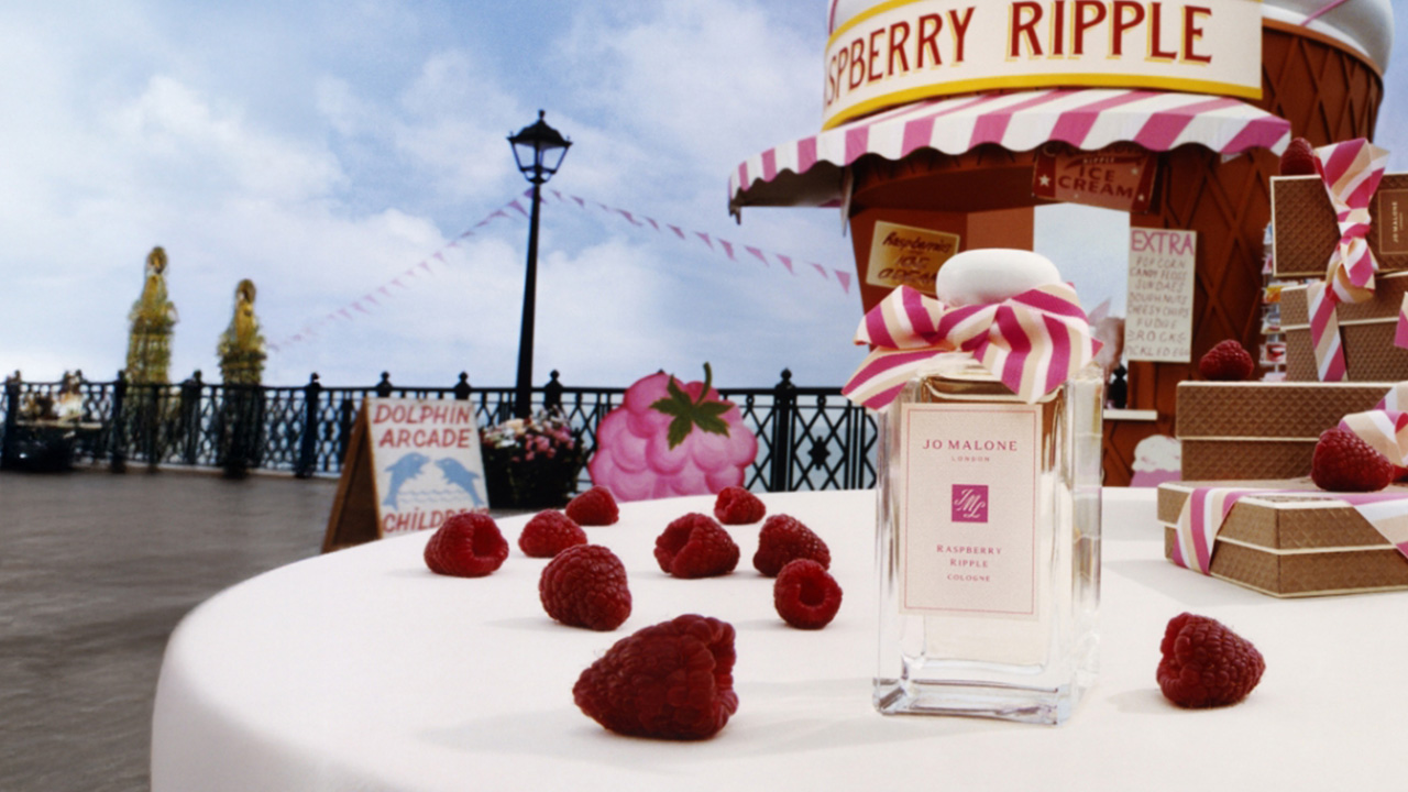Jo Malone London fragrance bottle surrounded by raspberries and ribbon, with a whimsical fairground backdrop.