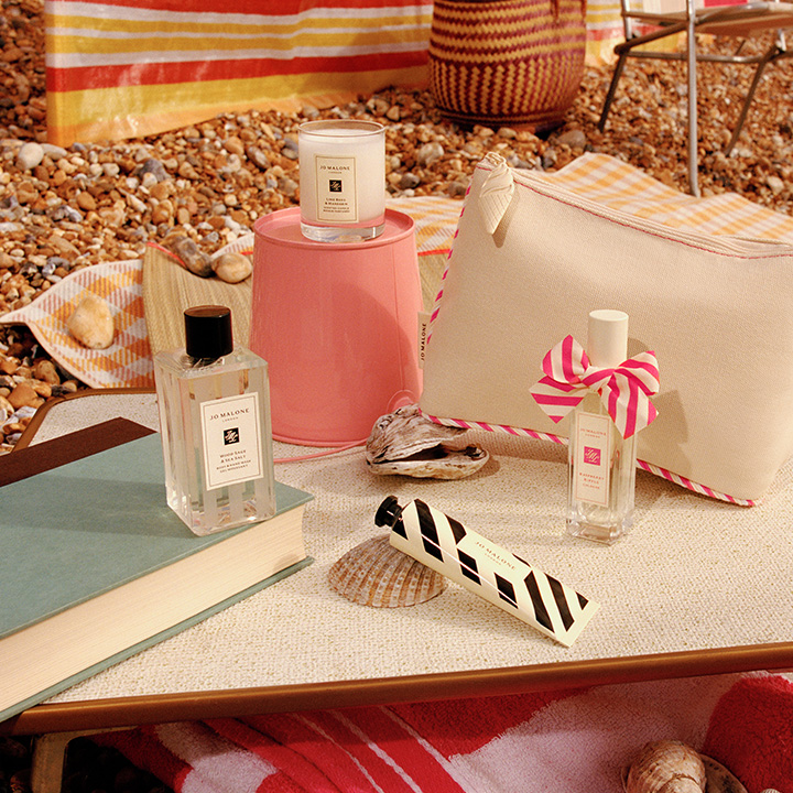 Jo Malone London fragrance bottles on a beach setup with striped towels, a bag, and seashells, creating a serene coastal atmosphere.