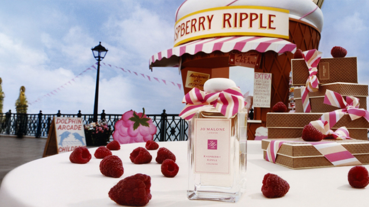 Jo Malone London fragrance bottle surrounded by raspberries and ribbon, with a whimsical fairground backdrop.