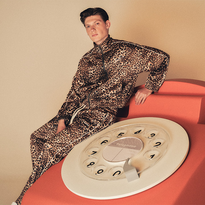 A man in a leopard-print outfit leans on a large, red rotary phone sculpture.