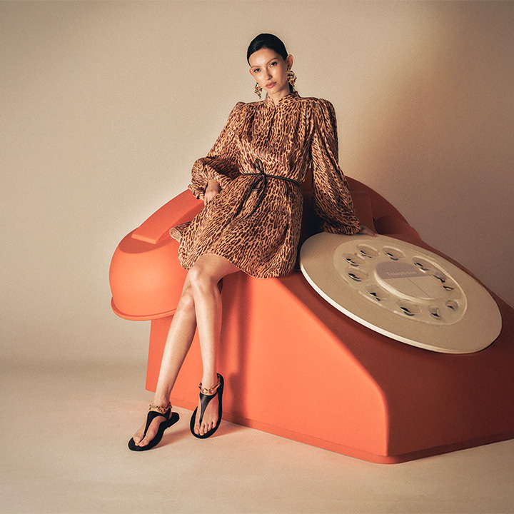 A woman in a patterned dress sits on a large, orange, vintage-style rotary phone.