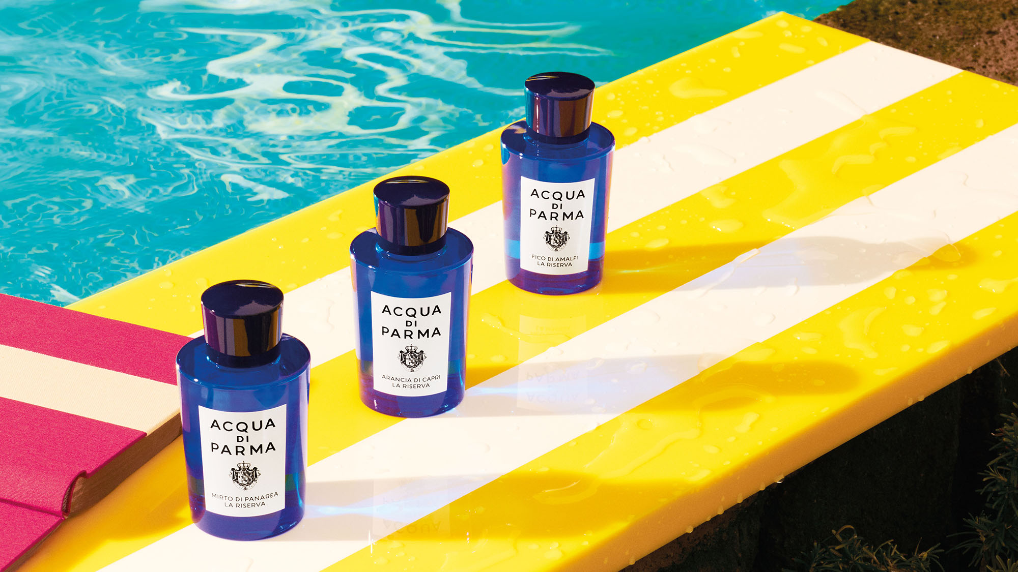 Three Acqua di Parma bottles are placed on a yellow and white striped poolside board, next to a swimming pool.