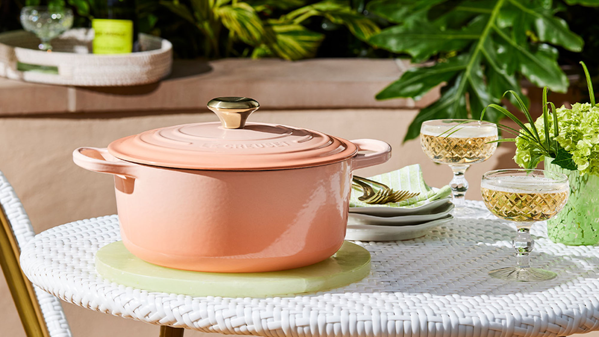 A pink Le Cresuet dish sits on a white wicker table with plates, forks, glasses, and a green floral arrangement, set in an outdoor garden scene.