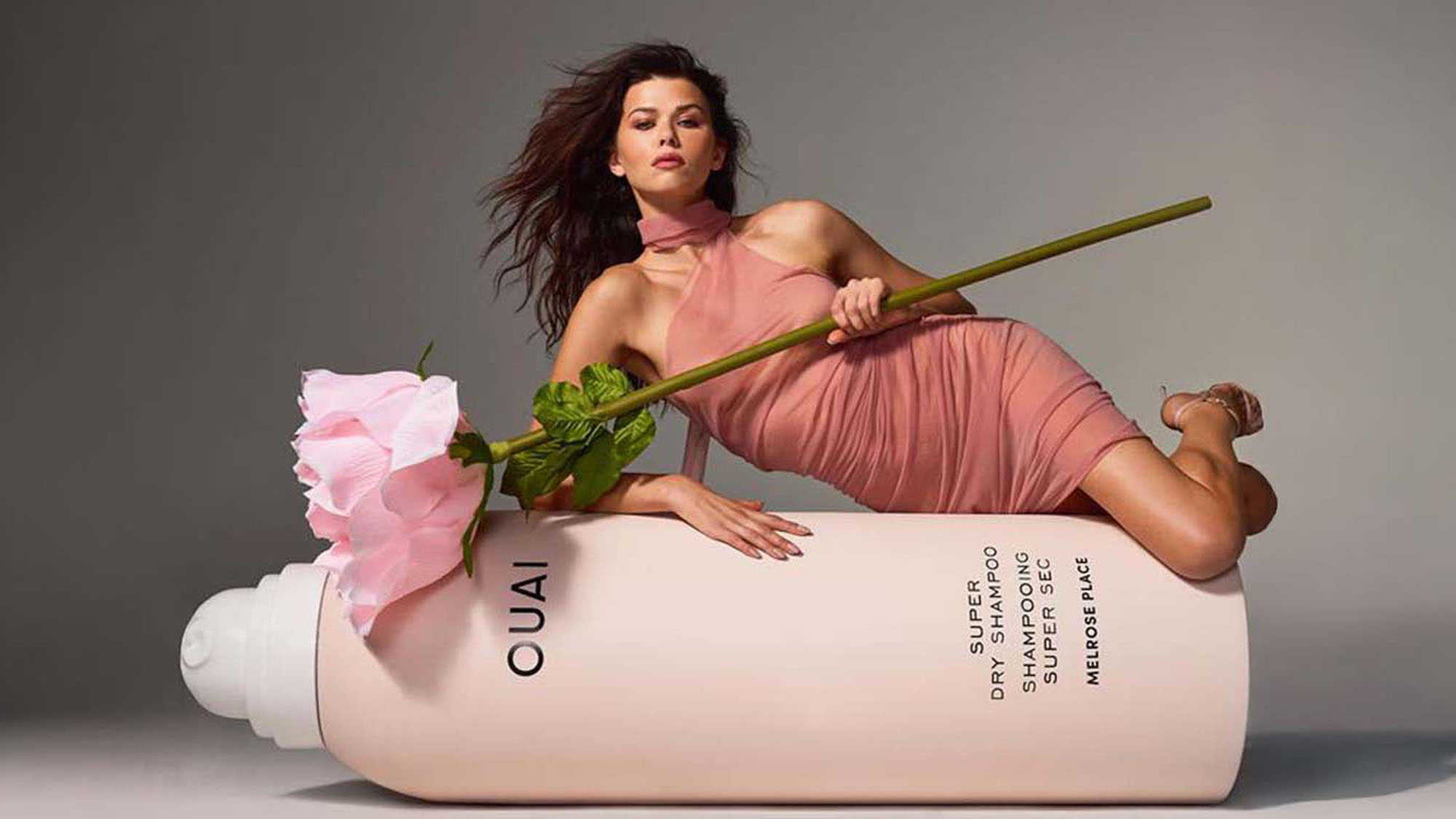 A woman in a pink dress reclines on a large shampoo bottle, holding a giant pink rose.
