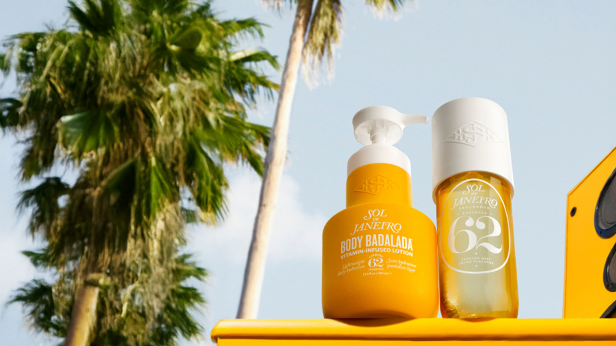 Two yellow Sol de Janeiro products are displayed outdoors with a palm tree and blue sky in the background.
