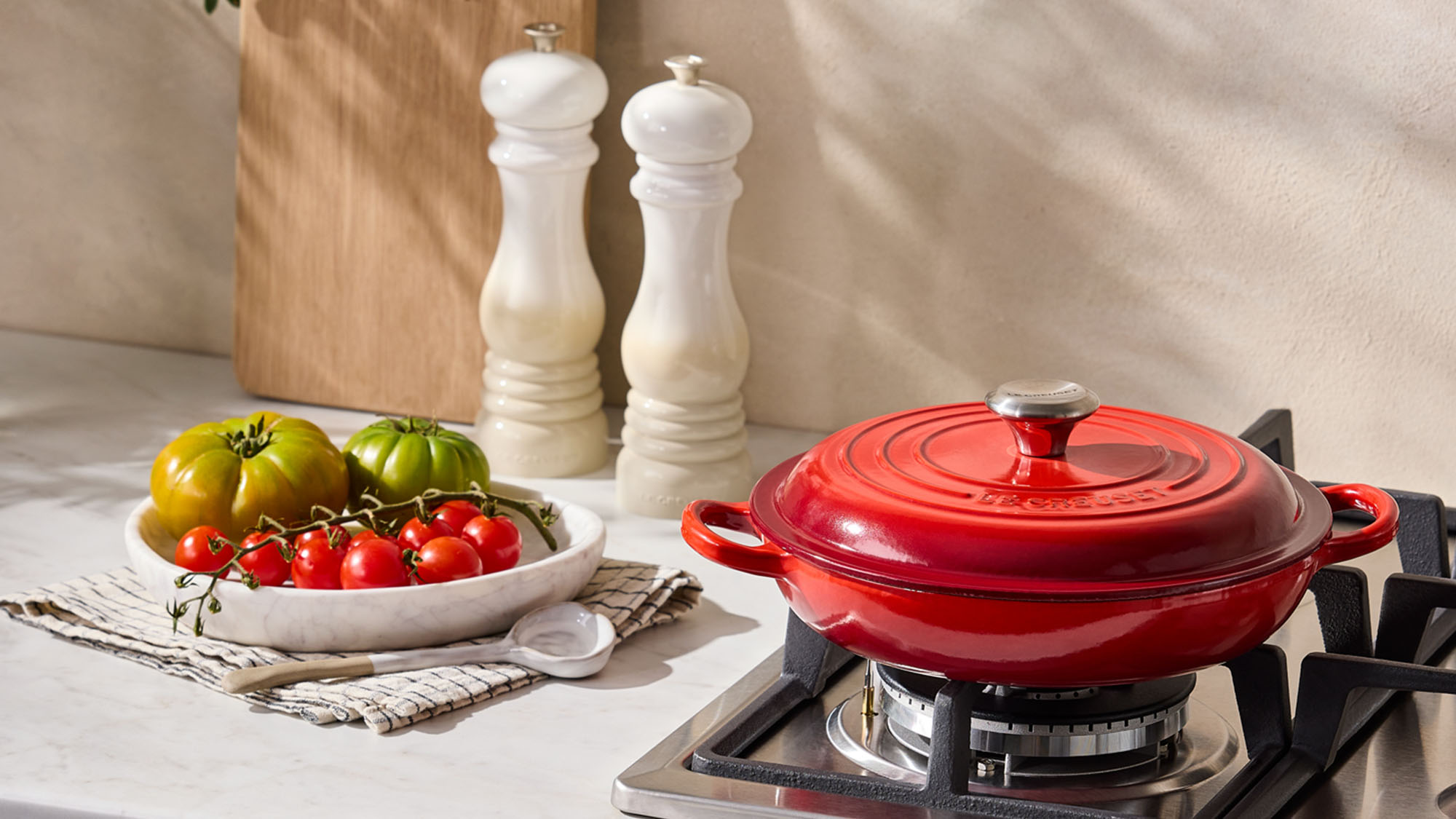 A kitchen scene with a red pot on a stove, a plate of tomatoes, salt and pepper shakers, and a cutting board with foliage.