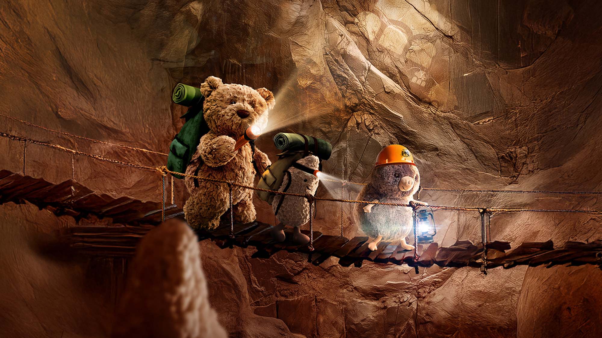 Three stuffed animals, including a bear and a mole, explore a cave with backpacks and lanterns on a wooden bridge.