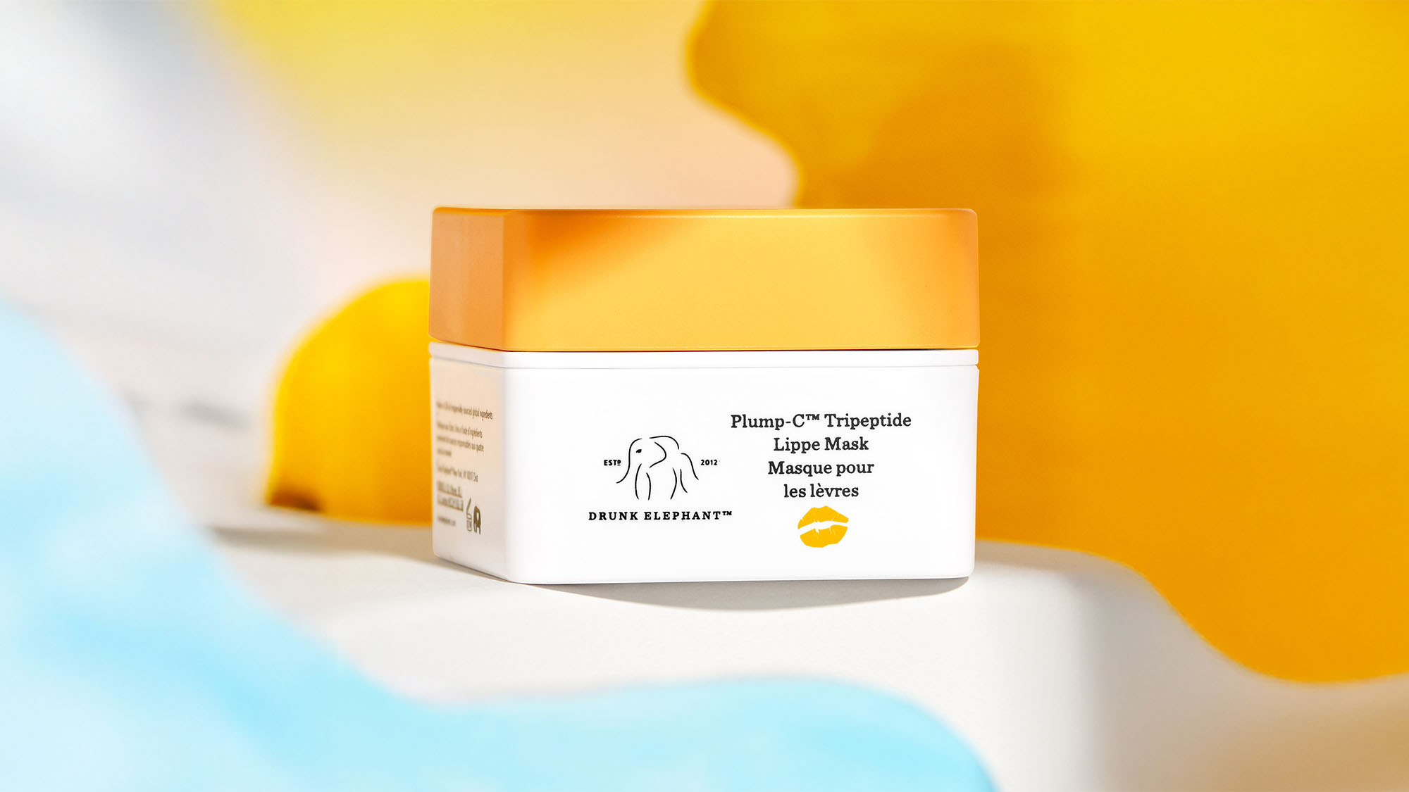 A Drunk Elephant lip mask with an orange lid and white container against a colorful background.