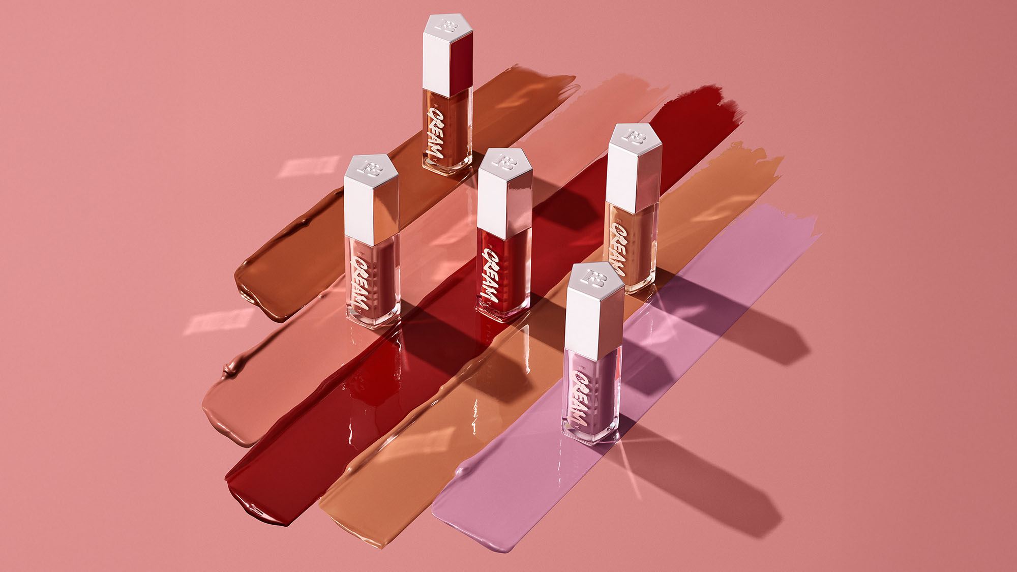Five lip gloss tubes on swatches of assorted shades on a pink background.