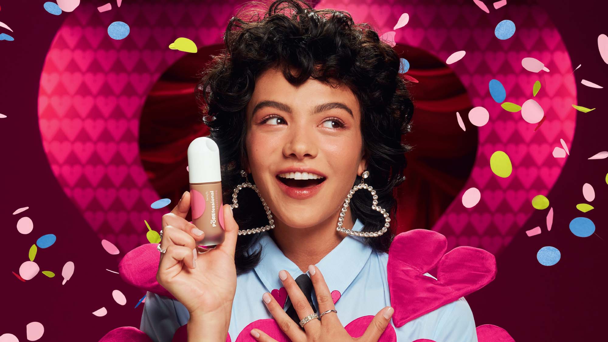 A person with heart-shaped earrings holds a makeup product surrounded by colorful confetti and pink hearts.