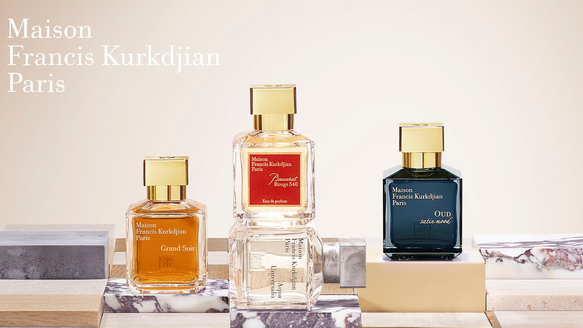 Three Maison Francis Kurkdjian perfume bottles are displayed on stacked marble slabs against a beige background.