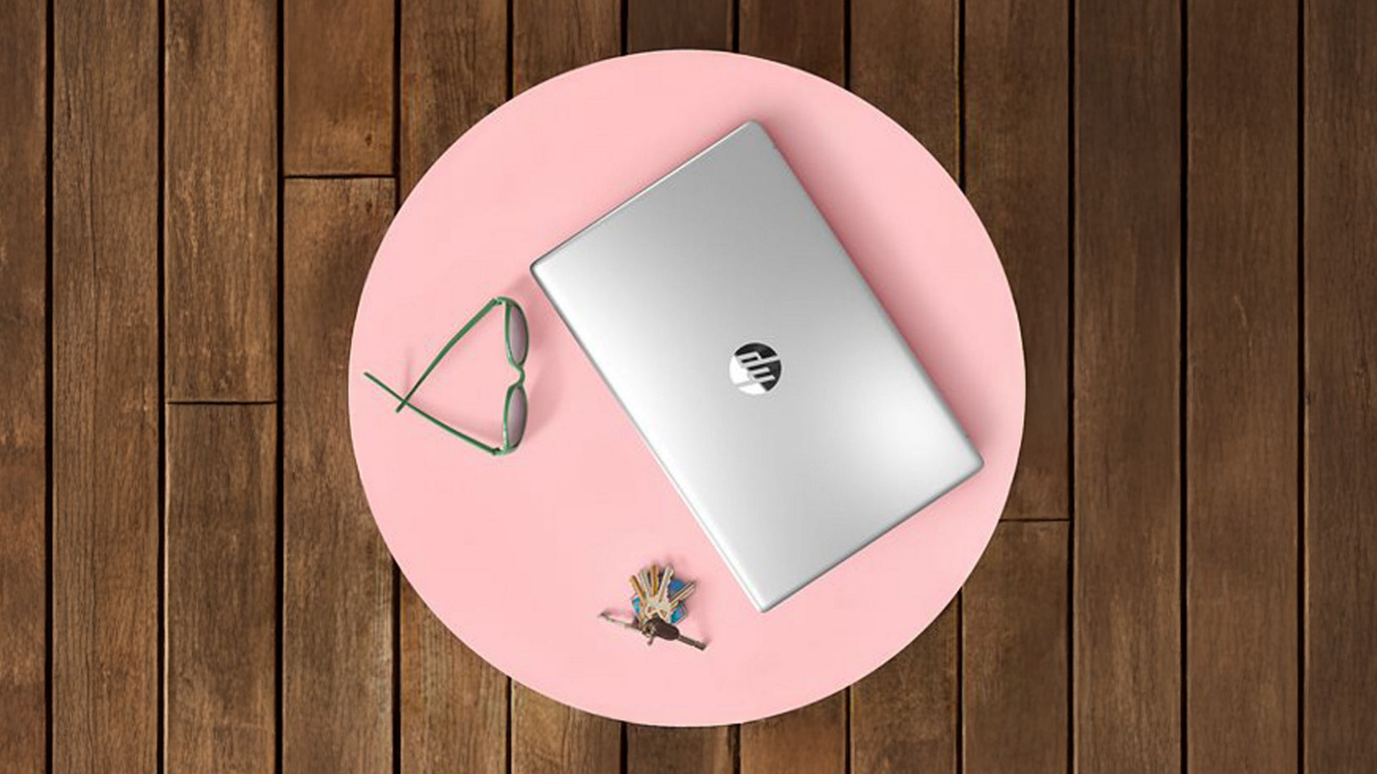 A closed silver laptop, sunglasses, and a small bouquet are arranged on a round pink table, set against a wooden floor.