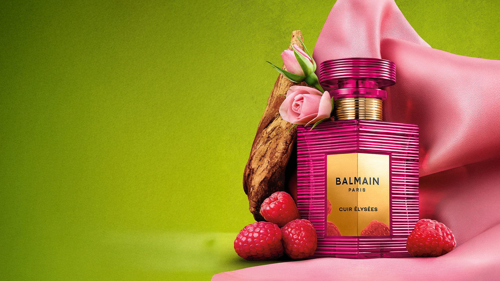A pink Balmain perfume bottle with a gold label is surrounded by raspberries, a rose, and a piece of wood, set against pink fabric and a green background.