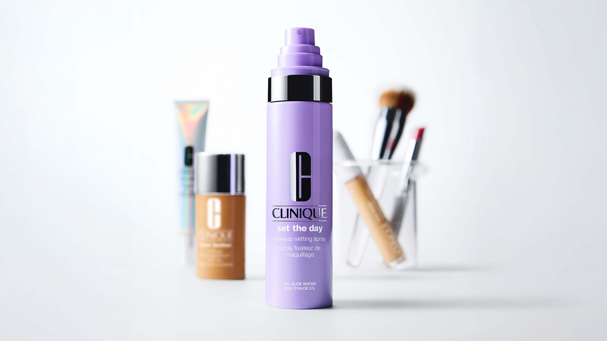 A purple Clinique setting spray bottle is in focus, with blurred makeup items in the background.