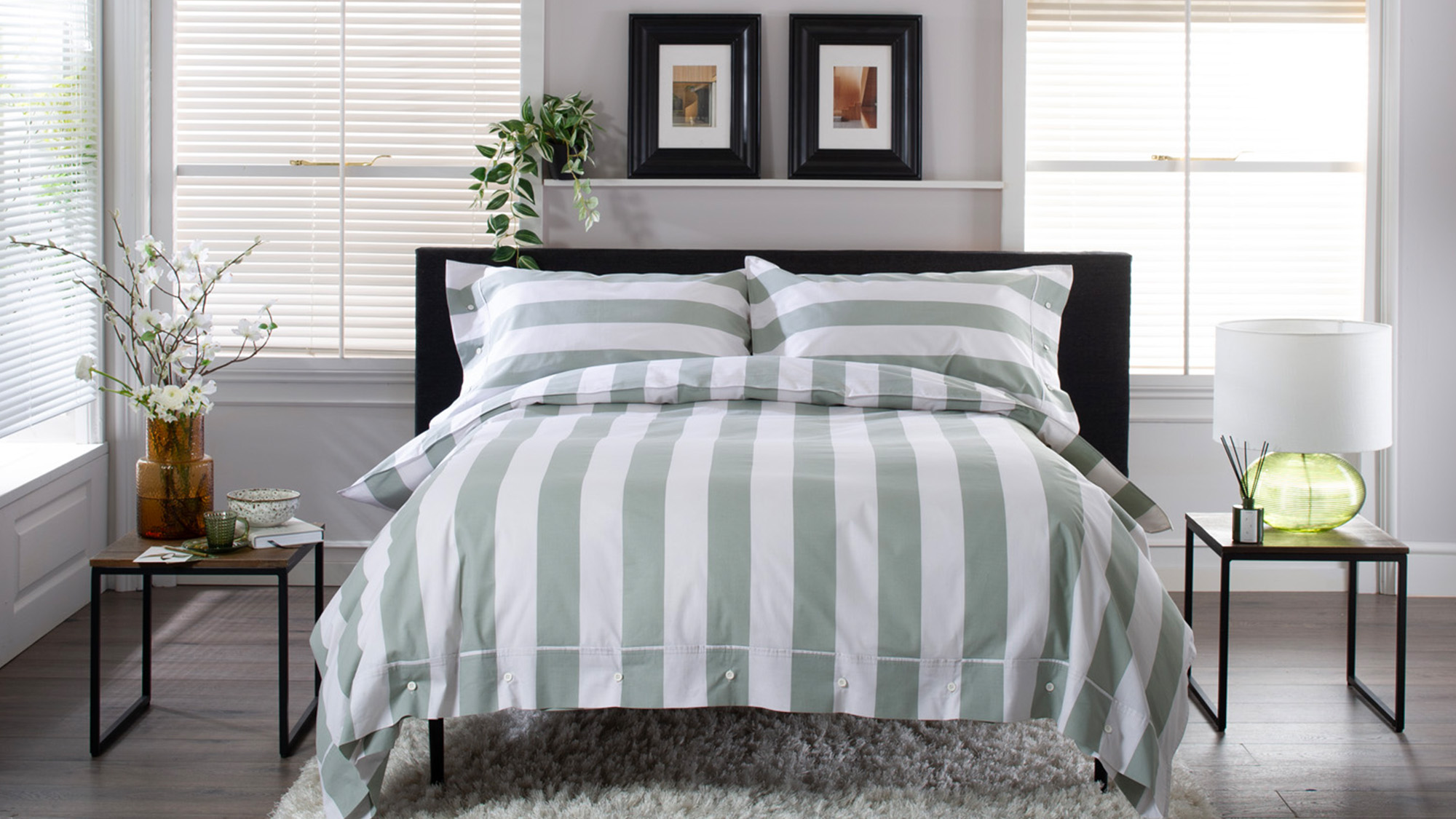 Modern bedroom with a striped bedspread, two side tables, and decorative plants. The walls have framed artwork above the bed.